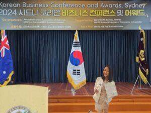 Elie Hoju in professional attire speaks at a podium, featuring the South Korean flag as a backdrop.