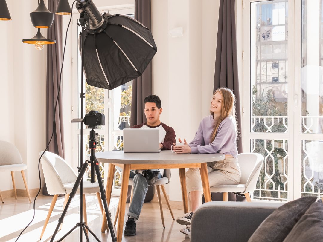 A man and woman sit at a table, focused on a laptop, engaged in discussion or collaboration.