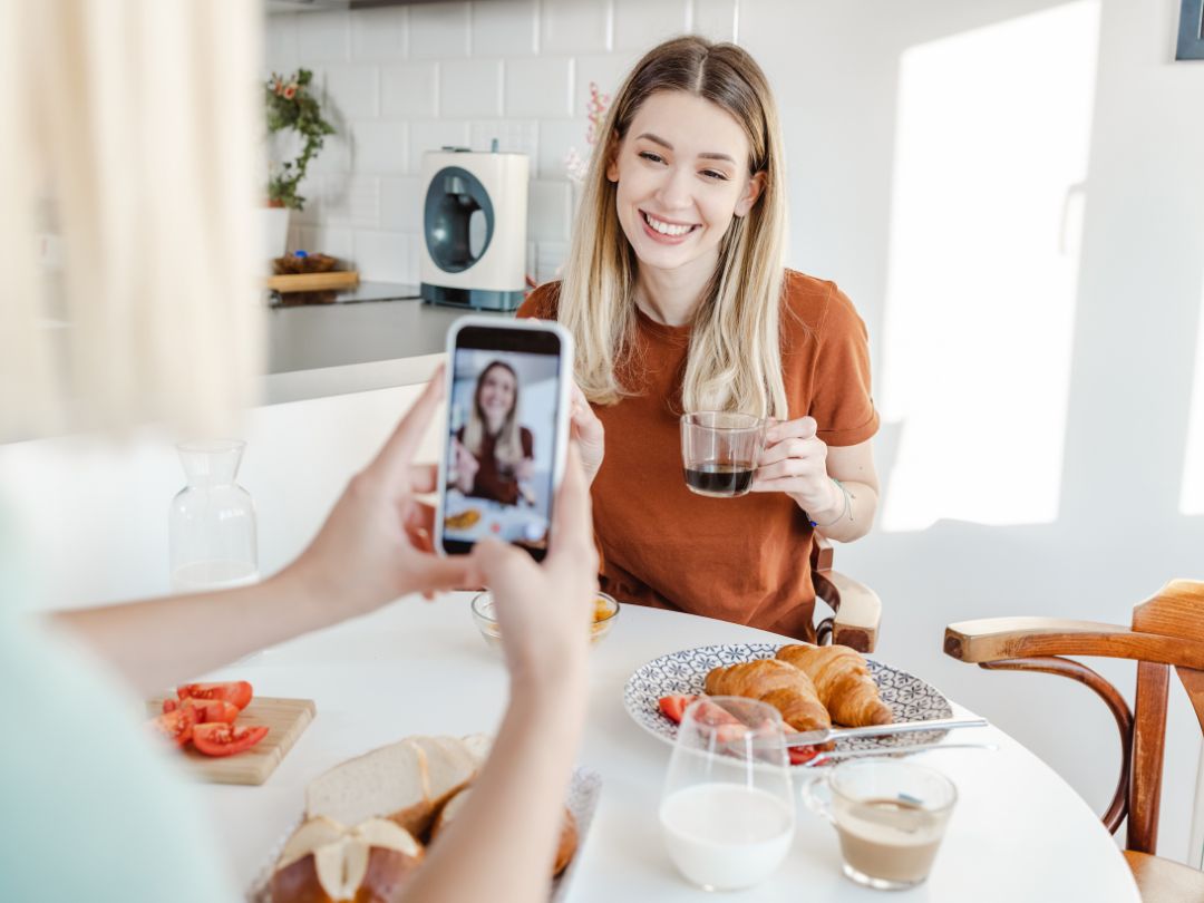 A woman sitting at a table takes a selfie with her phone, smiling and enjoying the moment.