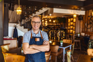 Proud, mature café owner with apron standing on the door of restaurant and looking at camera, smiling