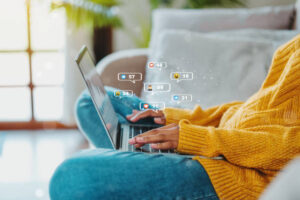 A woman on a couch uses her laptop, with various social media icons displayed around her, indicating online engagement.
